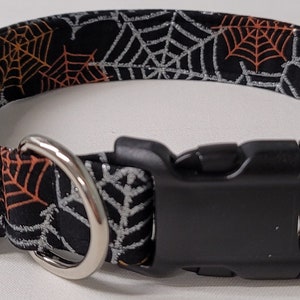 May include: Black dog collar with a silver and orange spiderweb pattern. The collar has a black plastic buckle and a silver D-ring.