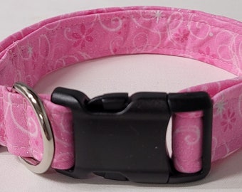 May include: A pink dog collar with a white floral pattern. The collar has a black plastic buckle and a silver metal D-ring. The fabric is a light pink with a repeating design of flowers and swirls.