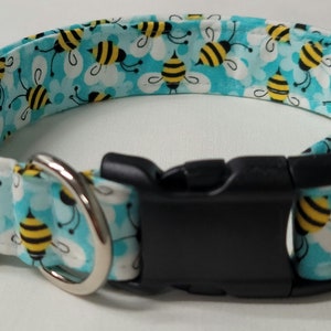 May include: A blue and white dog collar with a pattern of yellow and black bees. The collar has a black plastic buckle and a silver metal D-ring.