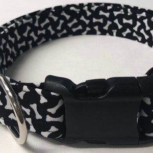 May include: Black and white dog collar with a bone pattern. The collar has a black plastic buckle and a silver D-ring.