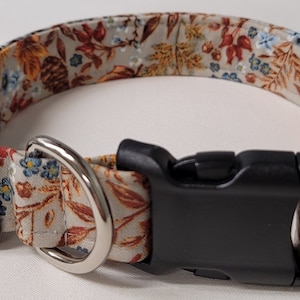 May include: A beige dog collar with a floral pattern featuring brown leaves, pine cones, and blue flowers. The collar has a black buckle and a silver D-ring.