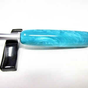 May include: A teal and white marbled pen with a silver tip and accents rests on a black pen holder. The pen has a rounded shape and a smooth, glossy finish. The pen holder is rectangular with a curved design.