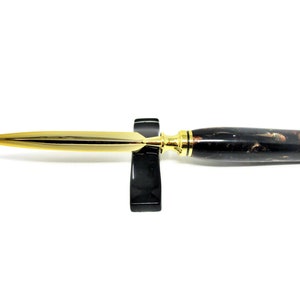 Könnte beinhalten: Ein goldener Brieföffner mit einem schwarz-braunen marmorierten Griff. Der Brieföffner steht auf einem schwarzen Ständer.