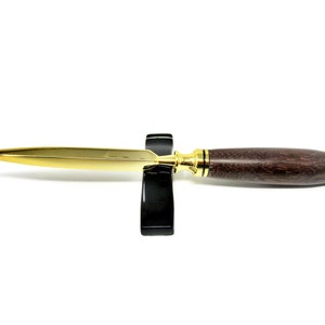 Könnte beinhalten: Ein goldener Brieföffner mit einem dunkelbraunen Griff und einer goldenen Spitze. Der Brieföffner liegt auf einem schwarzen Ständer.