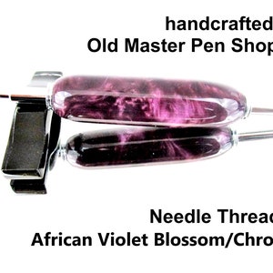 May include: A handcrafted needle threader with an African Violet Blossom design and chrome accents. The tool is displayed on a reflective surface, showcasing its purple and white marbled pattern. The text "handcrafted by Old Master Pen Shoppe" is visible.