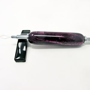 May include: A purple and silver beading tool with a diamond-shaped wire tip. The tool has a cylindrical handle with a marbled purple design and is resting on a black stand. The tool is designed for crafting and jewelry making.