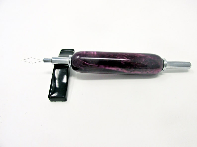 May include: A purple and silver beading tool with a diamond-shaped wire tip. The tool has a cylindrical handle with a marbled purple design and is resting on a black stand. The tool is designed for crafting and jewelry making.