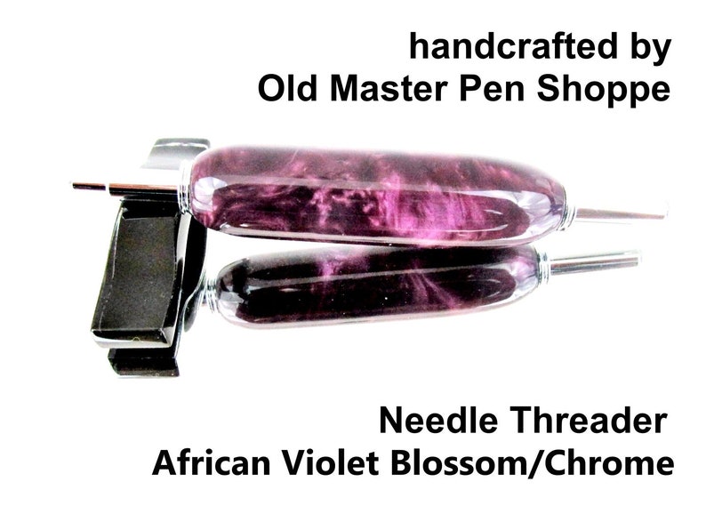 May include: A handcrafted needle threader with an African Violet Blossom design and chrome accents. The pen-like tool features a vibrant purple and pink marbled pattern. The text "handcrafted by Old Master Pen Shoppe" is visible.