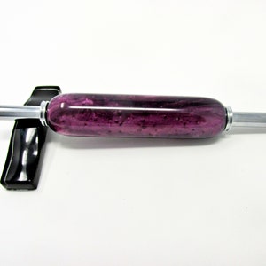 May include: A rolling pin with a vibrant purple and black speckled design. The cylindrical body is accented with silver-colored metal handles. The rolling pin rests on a black stand, set against a white background.
