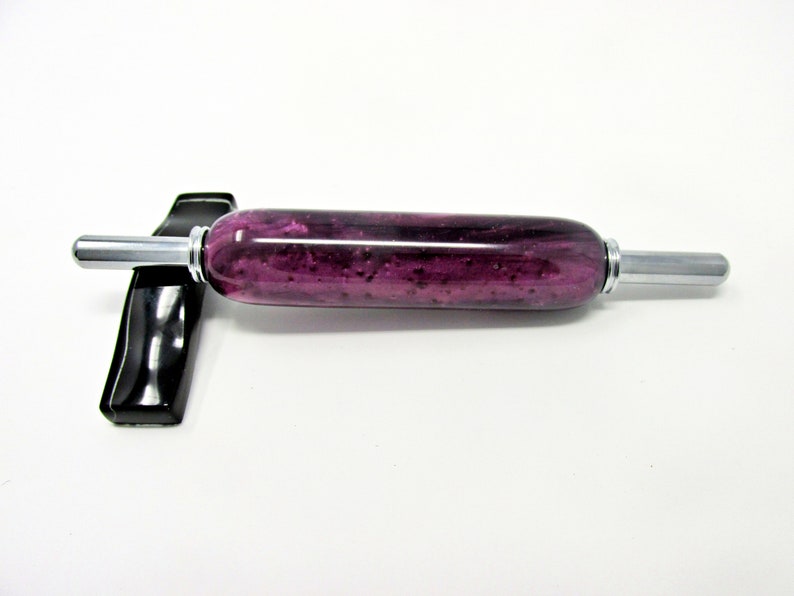May include: A rolling pin with a vibrant purple and black speckled design. The cylindrical body is accented with silver-colored metal handles. The rolling pin rests on a black stand, set against a white background.