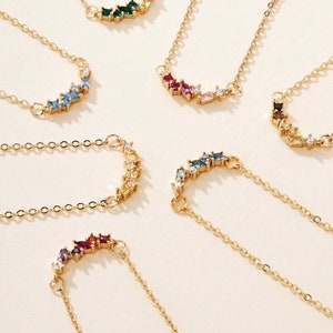 Könnte beinhalten: Ein Set aus sieben goldenen Halsketten mit einem Halbmond-Design, das mit verschiedenen farbigen Edelsteinen besetzt ist. Die Halsketten sind in einem Halbkreis auf weißem Hintergrund angeordnet.