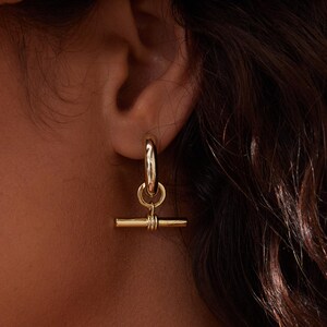 May include: A gold-coloured hoop earring with a horizontal bar pendant. The bar is connected to the hoop with a small, decorative loop. The earring is a simple, yet elegant piece of jewellery.