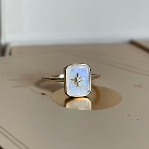 May include: A gold ring with a rectangular moonstone setting featuring a small diamond in the centre, forming a star shape.