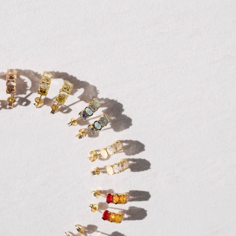 May include: A set of gold hoop earrings with different colored gemstones. The earrings are arranged in a rainbow pattern, starting with red and ending with blue.