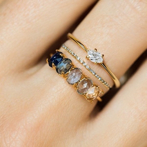 Puede incluir: Una colección de anillos de oro adornados con piedras preciosas. Los anillos presentan un diamante talla marquesa y una fila de gemas de corte ovalado en tonos azules, grises y melocotón. Los anillos están apilados en un dedo.