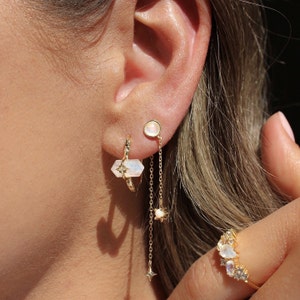 May include: Gold earrings with moonstone and star accents. The earrings are a hoop style with a dangling chain featuring a star charm. A separate moonstone stud earring is also shown.