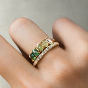 Puede incluir: Una mano muestra tres anillos de oro. El primer anillo presenta una fila de gemas verdes vibrantes. El segundo anillo tiene una fila de gemas de color verde claro. El tercer anillo es una banda de oro con pequeñas gemas claras.