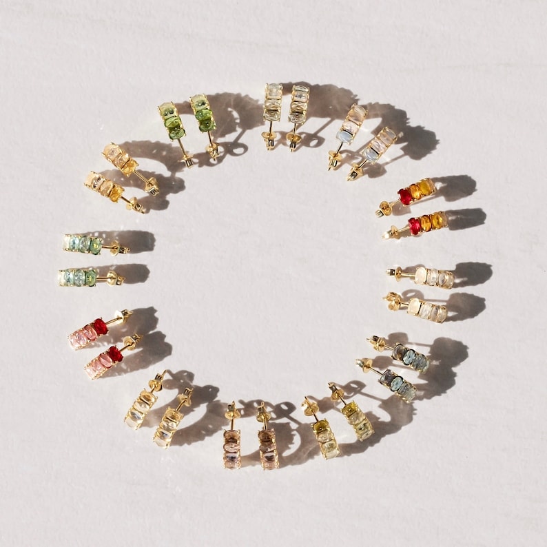 May include: A set of 12 pairs of gold hoop earrings with colorful gemstones. Each pair features a different color combination of gemstones, creating a rainbow effect.