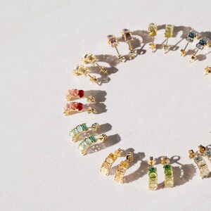 May include: A set of rainbow colored gemstone hoop earrings. The earrings are made of gold and feature a variety of gemstones, including pink, red, green, blue, and yellow. The earrings are arranged in a circle on a white background.