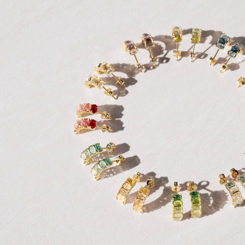 May include: A set of rainbow colored gemstone hoop earrings. The earrings are made of gold and feature a variety of gemstones, including pink, red, green, blue, and yellow. The earrings are arranged in a circle on a white background.