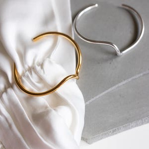 Può includere: Due bracciali aperti, uno in tonalità oro lucido e uno in tonalità argento lucido. Ogni bracciale presenta una fascia metallica liscia e continua con una distinta curva a forma di onda al centro, creando un design aperto per il polso.