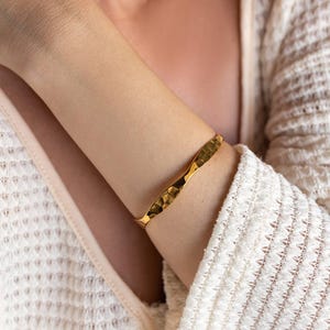 Puede incluir: Un brazalete rígido de metal en tono dorado que presenta una silueta ondulada y una superficie martillada y texturizada que refleja la luz sobre su acabado pulido, creando una serie de reflejos brillantes y sombras profundas.