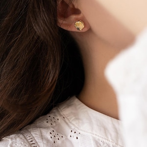 Peut inclure: Une petite boucle d'oreille à clou en or en forme de coquillage avec des stries verticales. La boucle d'oreille est fixée au lobe, présentant une finition métallique polie qui reflète la lumière, créant un bijou simple et distinctif.