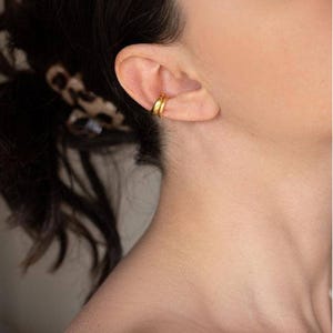 Puede incluir: Primer plano de un ear cuff dorado usado en la oreja. El ear cuff tiene un diseño simple y minimalista, con una superficie texturizada. La joya es de color oro brillante.