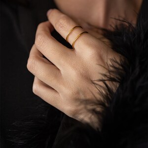 Puede incluir: Un conjunto de dos anillos de metal fino y liso en tono dorado que se llevan en un solo dedo. Los anillos presentan un diseño de banda circular simple con un acabado pulido, creando una apariencia apilada sobre un fondo de tela oscura y texturizada.