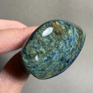 May include: A polished, oval-shaped gemstone with swirling patterns of blue, green, and gold. The stone has a glossy finish and is held in a hand, showcasing its natural beauty and unique color variations. The gemstone is a decorative item.