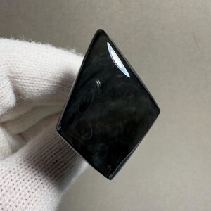 K&ouml;nnte beinhalten: Ein polierter, schwarzer Obsidian-Edelstein in Diamantenform. Die Oberfl&auml;che reflektiert Licht und zeigt wirbelnde Muster im Stein. Der Edelstein wird vor einem wei&szlig;en Hintergrund gehalten.