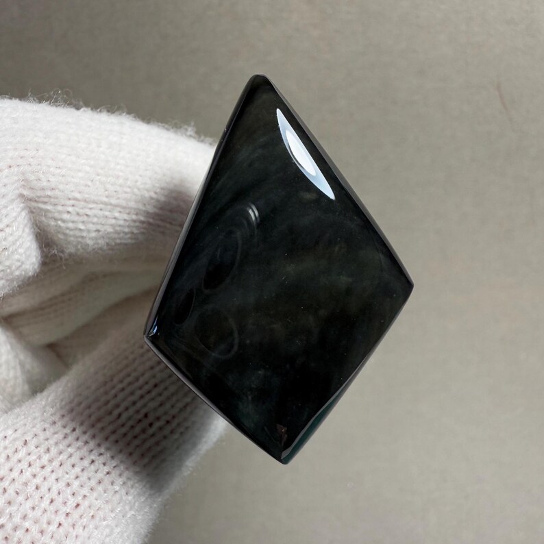 K&ouml;nnte beinhalten: Ein polierter, schwarzer Obsidian-Edelstein in Diamantenform. Die Oberfl&auml;che reflektiert Licht und zeigt wirbelnde Muster im Stein. Der Edelstein wird vor einem wei&szlig;en Hintergrund gehalten.
