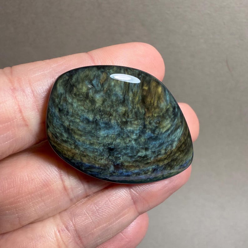 May include: A polished, oval-shaped gemstone with a swirling pattern of deep blues, greens, and hints of gold. The stone is held in a hand, showcasing its smooth, reflective surface. The gemstone's unique color variations create a captivating visual texture.
