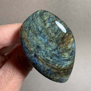 May include: A polished, oval-shaped stone with a swirling pattern of blue, green, and gold hues. The stone has a glossy finish and is held in a hand, showcasing its unique color variations and texture.