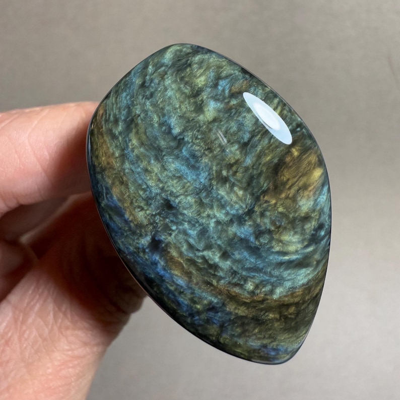 May include: A polished, oval-shaped stone with a swirling pattern of blue, green, and gold hues. The stone has a glossy finish and is held in a hand, showcasing its unique color variations and texture.