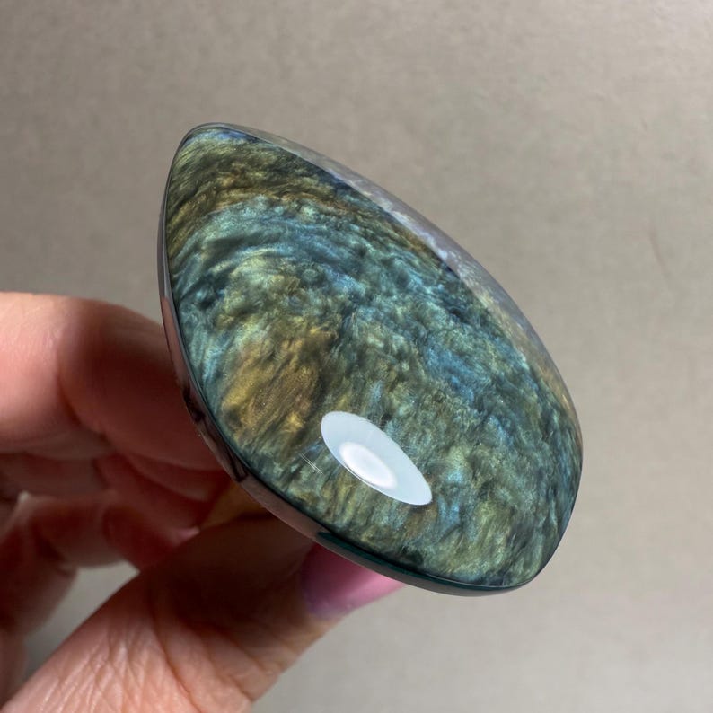 May include: A teardrop-shaped, polished gemstone with a mix of blue, green, and gold colors. The stone has a glossy, reflective surface and a dark border. The gemstone is held in a hand.