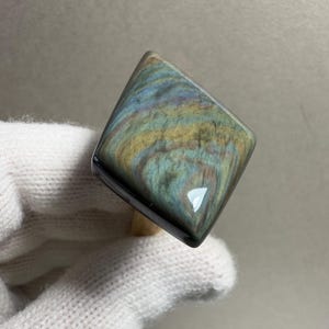 K&ouml;nnte beinhalten: Ein quadratischer Flaschenverschluss mit polierter, schillernder Oberfl&auml;che. Er zeigt wirbelnde Muster in Blau-, Gr&uuml;n- und Goldt&ouml;nen. Die Basis ist dunkel und an einem Holzkorken befestigt. Ein dekorativer Artikel f&uuml;r Flaschen.