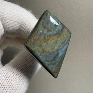 K&ouml;nnte beinhalten: Ein polierter, rechteckiger Edelstein mit einem einzigartigen, wirbelnden Muster aus Gr&uuml;n, Blau und Gold. Der Stein hat eine gl&auml;nzende Oberfl&auml;che und einen dunklen Rand. Der Edelstein wird in einem wei&szlig;en Handschuh gehalten.