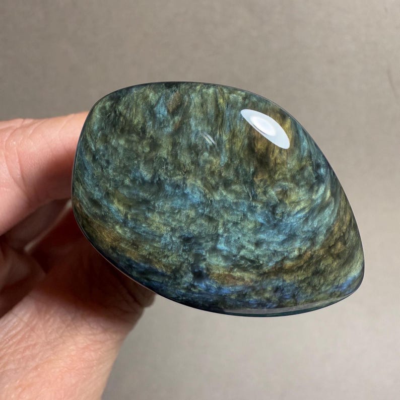 May include: A polished, oval-shaped gemstone with a swirling pattern of blues, greens, and golds. The stone has a glossy, reflective surface, and is held in a hand.