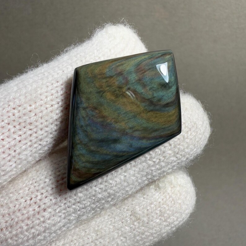 K&ouml;nnte beinhalten: Ein polierter, schillernder Obsidian-Edelstein mit einer einzigartigen, unregelm&auml;&szlig;igen Form. Der Stein zeigt wirbelnde Muster in Gr&uuml;n-, Blau- und Goldt&ouml;nen. Der Obsidian wird in einem wei&szlig;en Handschuh gehalten, was die reflektierende Oberfl&auml;che hervorhebt.
