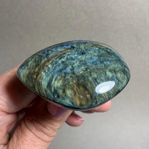 May include: A polished, teardrop-shaped gemstone with a swirling pattern of blue, green, and gold hues. The stone is held in a hand, showcasing its smooth, reflective surface and natural color variations. The gemstone is a unique, eye-catching piece.