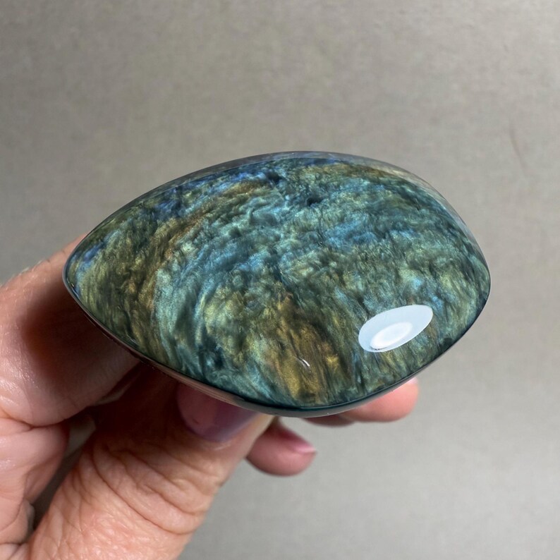 May include: A polished, teardrop-shaped gemstone with a swirling pattern of blue, green, and gold hues. The stone is held in a hand, showcasing its smooth, reflective surface and natural color variations. The gemstone is a unique, eye-catching piece.