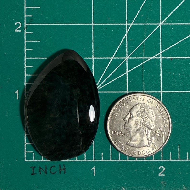 May include: A polished, teardrop-shaped gemstone in shades of dark green and black, next to a U.S. quarter for size comparison. The gemstone is approximately 1 inch long.