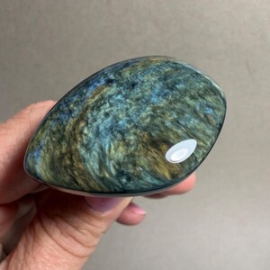 May include: A polished, oval-shaped gemstone with a swirling pattern of blue, green, and gold hues. The stone is held in a hand, showcasing its smooth, reflective surface and unique color variations.