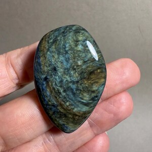 May include: A polished, oval-shaped gemstone with a swirling pattern of blue, green, and gold hues. The stone has a smooth, glossy surface and a natural, organic appearance. The gemstone is held in a hand.