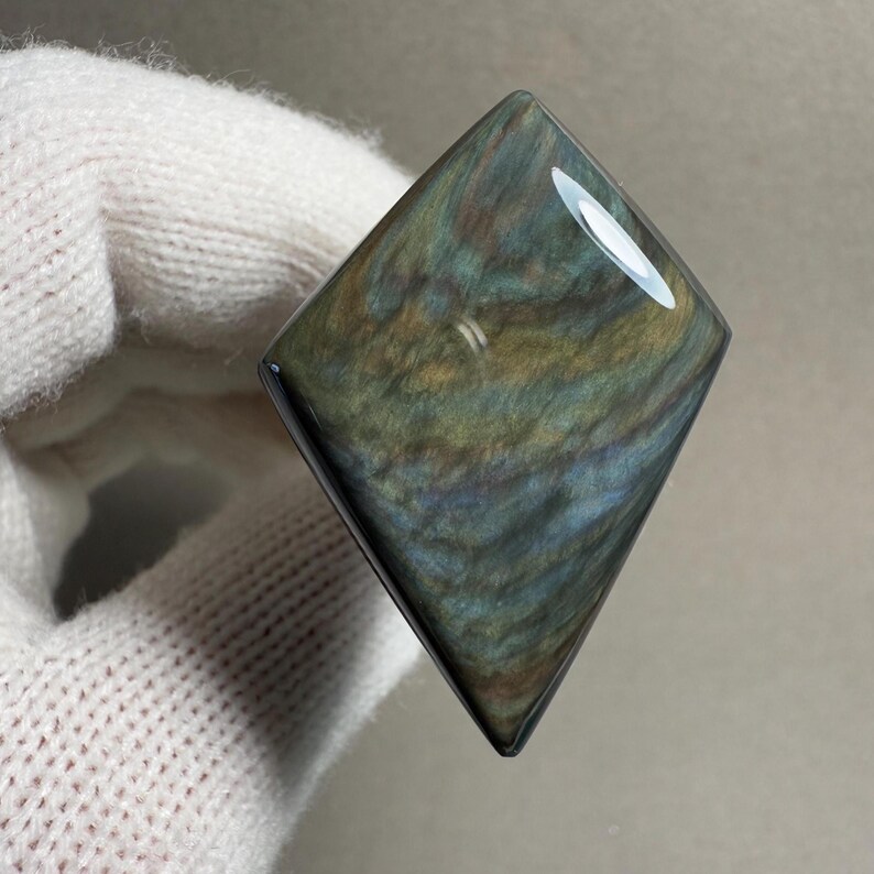 K&ouml;nnte beinhalten: Ein polierter, irisierender Obsidian-Edelstein in Diamantenform. Der Stein zeigt wirbelnde Muster in Blau, Gr&uuml;n und Gold. Der Edelstein wird in einem wei&szlig;en Handschuh gehalten.