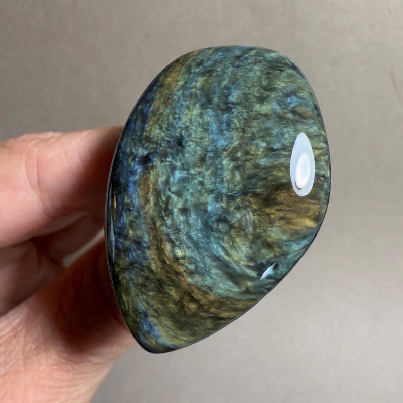 May include: A polished, oval-shaped stone with swirling patterns of blue, green, and gold. The stone has a glossy finish and a smooth texture. The object is held in a hand.