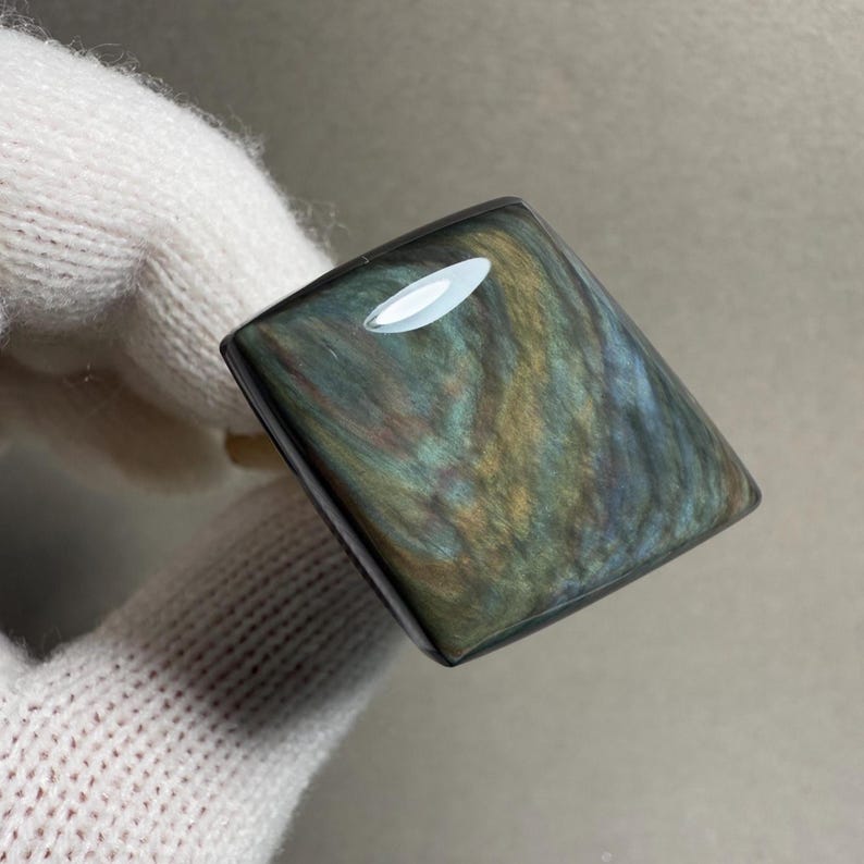 K&ouml;nnte beinhalten: Ein rechteckiger, polierter Obsidian-Edelstein mit schillernden Wirbeln aus Gold, Blau und Gr&uuml;n. Der Stein wird in einer behandschuhten Hand gehalten, die seine reflektierende Oberfl&auml;che und nat&uuml;rlichen Muster zeigt. Der Edelstein ist ein einzigartiges Schmuckst&uuml;ck.