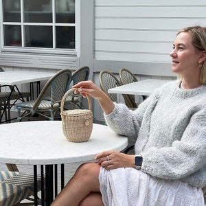 Può includere: Una donna con un vestito bianco e un maglione grigio chiaro è seduta a un tavolo con un piano in marmo bianco. Tiene una piccola borsa a cestello intrecciata. Il tavolo è circondato da sedie con cuscini a strisce blu e bianche. La donna indossa sandali marroni.