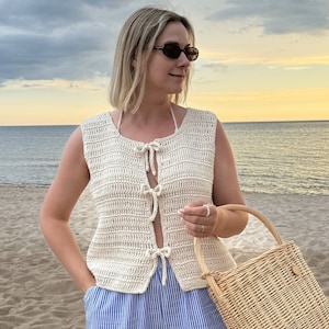 May include: A woman wearing a white crocheted sleeveless top with a tie at the front, blue and white striped pants, and sunglasses. She is holding a woven straw tote bag.